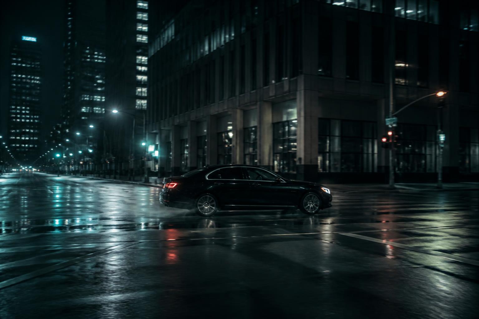 A black sedan crossing an empty city intersection at night, street lights reflected on wet asphalt.